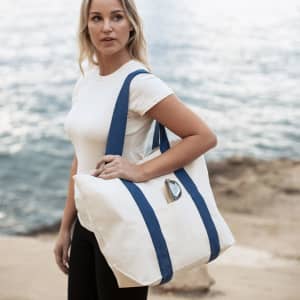 A tote bag in cream with navy blue straps is held by a person near a water background. The bag features a logo.