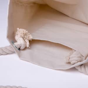 A cream tote bag featuring a woven handle and decorative knots at the base. It has a plain interior with no patterns.