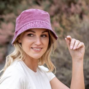 A pink bucket hat with a branding logo, worn by a woman against a natural background.