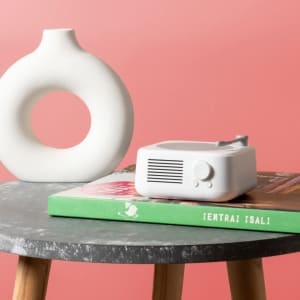 A compact white Bitels Speaker rests on a stack of books on a round, grey table, beside a ceramic vase.