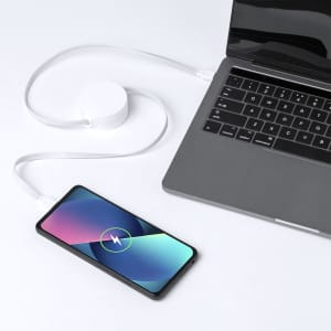 A white charging cable connects a smartphone to a laptop, on a minimalist surface. The phone displays a charging icon.