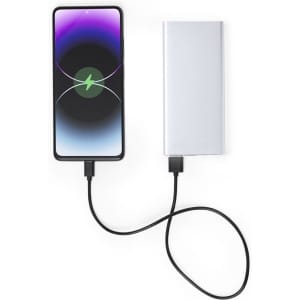 A silver power bank connected to a smartphone via a black charging cable. The phone displays a charging icon.