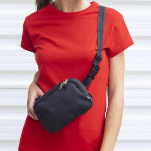 A black shoulder bag waistbag worn across a person's body, complemented by a red t-shirt. The bag features a zip closure and an adjustable strap.