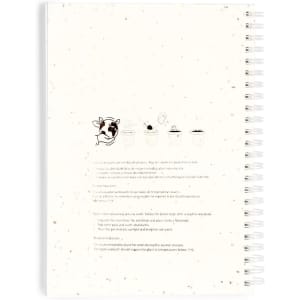 A spiral-bound notebook with a speckled cover featuring a cow illustration and a logo, in primarily white tones.