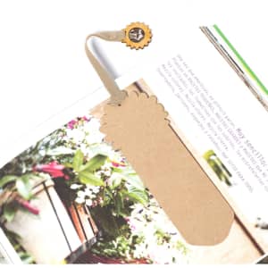 A brown paper bookmark with a decorative edge and a circular charm, placed in an open book among flowers.