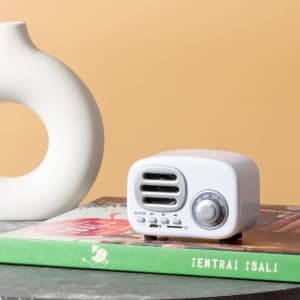 A white speaker with a vintage design sits on top of a green book next to a white vase against a warm background.