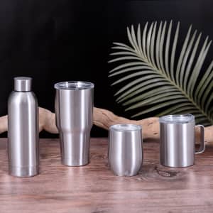 A drink bottle and three silver insulated cups on a wooden surface, featuring a logo. Background includes a green palm leaf.