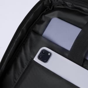 A grey laptop and a smartphone are placed inside a black backpack compartment. The backpack features a logo.