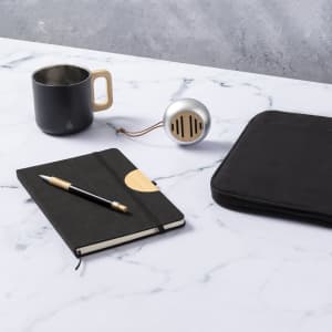 A black notebook with a pen, a black mug, a silver speaker, and a black laptop sleeve on a marble surface.