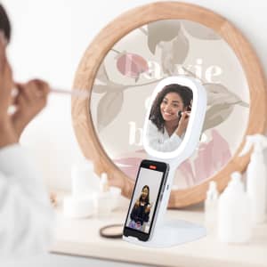 A multifunction mirror in white, featuring a phone holder, reflects a person applying makeup. The background includes floral decor.