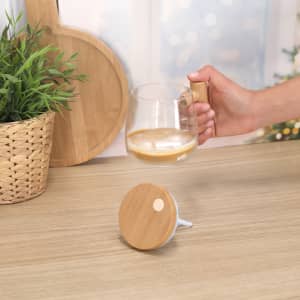 A transparent coffee mug with a wooden handle, set against a wooden surface and greenery in the background.