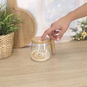 A coffee mug with a glass body and wooden lid, featuring a handle, sits on a wooden table near greenery.