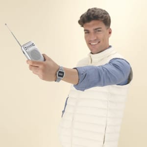 A man wearing a light vest holds a silver radio with a dial and antenna, smiling at the camera.