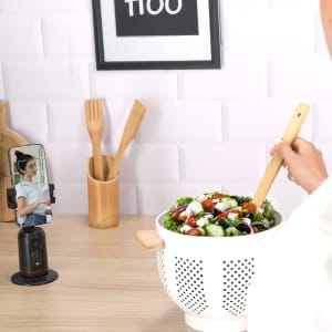 A black phone holder with a smartphone attached stands next to a bowl of salad on a wooden countertop.