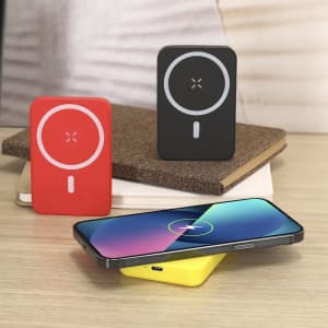 A red and a black power bank with circular logos are placed on a desk beside a smartphone resting on a yellow charger.