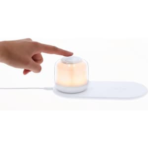 A wireless charger lamp featuring a white base and a transparent dome, emitting a warm glow, with a hand touching the top.