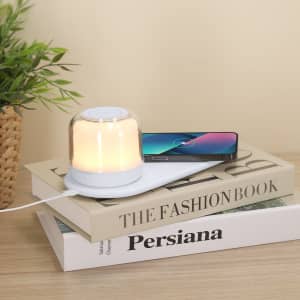 A wireless charger lamp in white with a soft glow, placed on stacked books next to a smartphone.