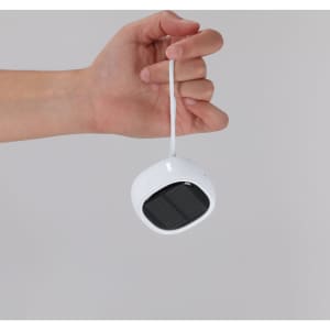 A hand holds a compact white speaker with a square solar panel on top and a hanging strap.