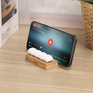 A wooden phone stand with white silicone grips, supporting a smartphone displaying a video.