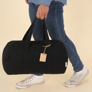 A person holds a black tote bag with handles, featuring a tag attached. The bag is made of sturdy material.