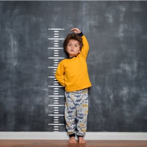 A child in a yellow top and patterned pants stands beside a height chart on a wall, raising their arm.