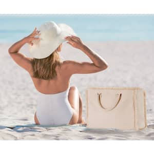 A cream tote bag with handles sits on the sand beside a woman wearing a white swimsuit and sunhat.