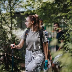 A woman in a grey shirt and light trousers holds a walking stick in a green, forested area. Two people are visible in the background.