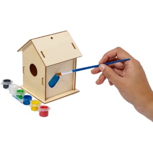 A wooden birdhouse in natural colour with a hand painting it blue, surrounded by paint pots in various colours.