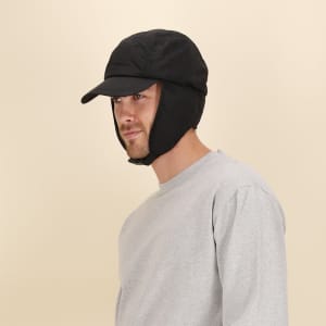A black cap with ear flaps worn by a model, featuring a rounded top and a peak. The model is in a grey sweatshirt.