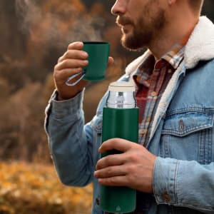 A drink bottle in a deep green colour being held by a man, with steam rising from a cup in his other hand.