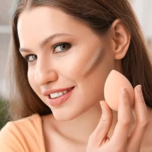 A beauty blender in a teardrop shape held by a smiling woman with long hair, showcasing its peach colour.