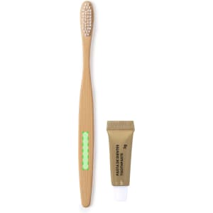 A bamboo toothbrush with a green accent alongside a small tube of toothpaste. Both items are positioned on a white background.