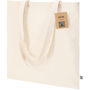 A cream-coloured tote bag with long handles and a tag. It features a logo and is made from Fairtrade materials.