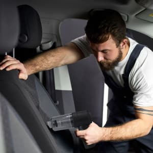 A vacuum cleaner in black with a transparent dust container, used for cleaning car seats.