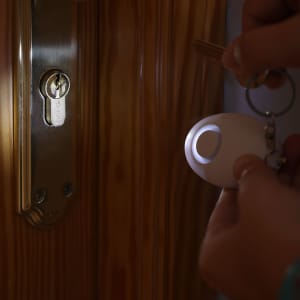 A white personal alarm in a person's hand, with a key in the background near a wooden door. The alarm has a small light.