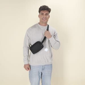 A person wearing a grey sweatshirt and light jeans holds a black sling bag, featuring a strap and a small tag.