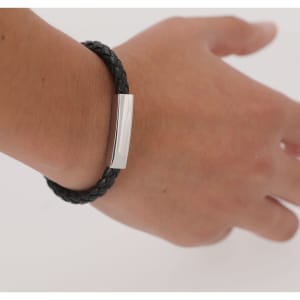 A black braided bracelet with a shiny silver clasp worn on a hand. The bracelet features a simple design.