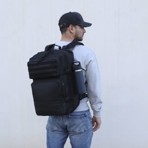 A black backpack with multiple compartments, featuring a logo, worn by a person with a drink bottle attached.