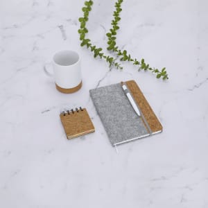 A grey notebook and a small notepad with a cork cover, alongside a white mug and a green plant on a marble surface.