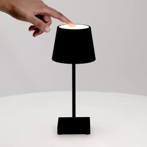 A black lamp with a cylindrical shade and a square base, being turned on by a hand.
