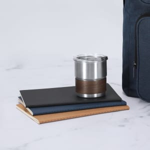 A stainless steel travel mug with a brown leather band sits on top of three notebooks, next to a dark blue bag.