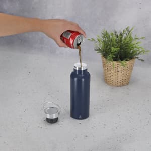 A drink bottle in navy blue being filled with a beverage, with a silver cap beside it and a plant in the background.