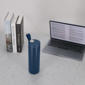 A drink bottle in deep blue, with a lid, placed on a desk beside books and a laptop. It features a logo.