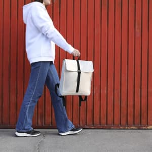 A woman carrying a beige Teylok backpack with black straps, walking against a red wall.