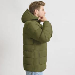 A green puffer jacket with a hood and zip pockets, worn by a model. The jacket features a quilted design.