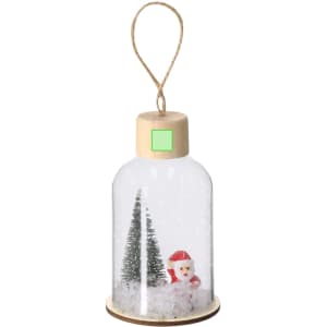 A decorative snow globe featuring a Santa figure, two green trees, and a snowy base, with a wooden top and hanging loop.