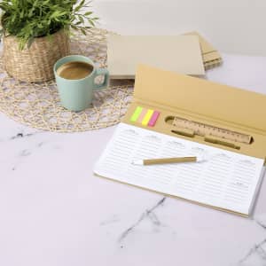 A yellow weekly planner open on a table, beside a cup of green, with sticky notes and a pencil, set on a marble surface.