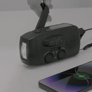 A black multifunction emergency radio with a hand crank and speaker, positioned next to a smartphone.