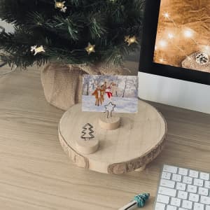 A wooden photo holder with two clips on a round base, featuring a festive scene against a backdrop of a computer and a Christmas tree.