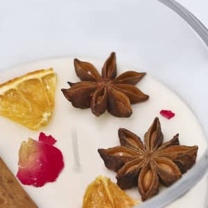 A candle featuring dried orange slices, star anise, and rose petals embedded in a creamy wax. The container is clear.
